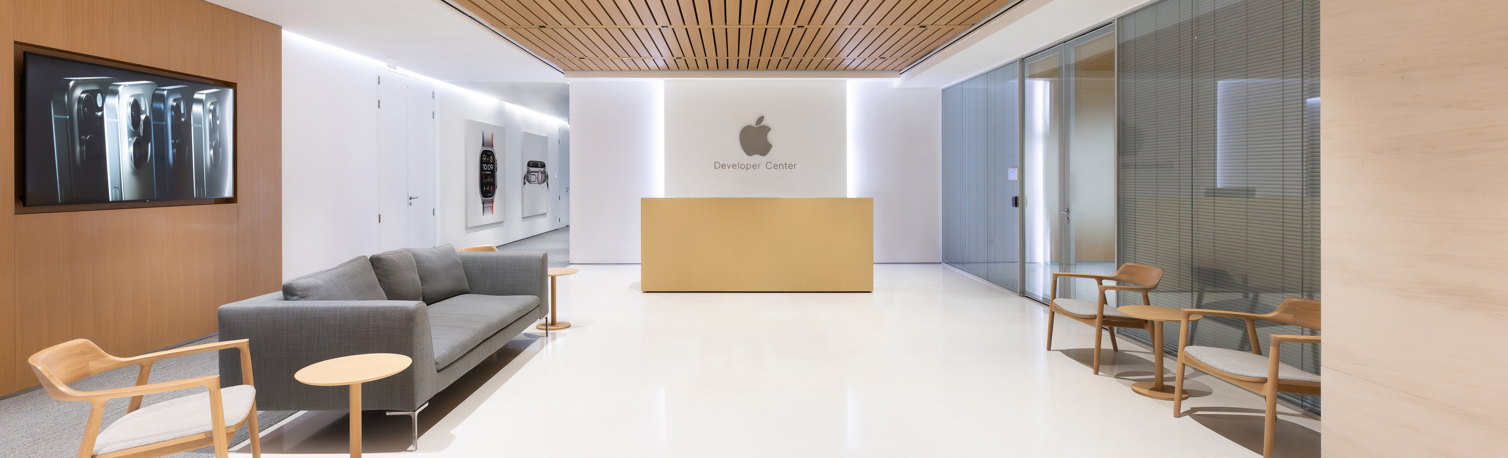 The interior of an Apple Developer Center, an open and brightly lit room with a gray couch, several wooden chairs, and a reception desk that sits underneath a large Apple logo.